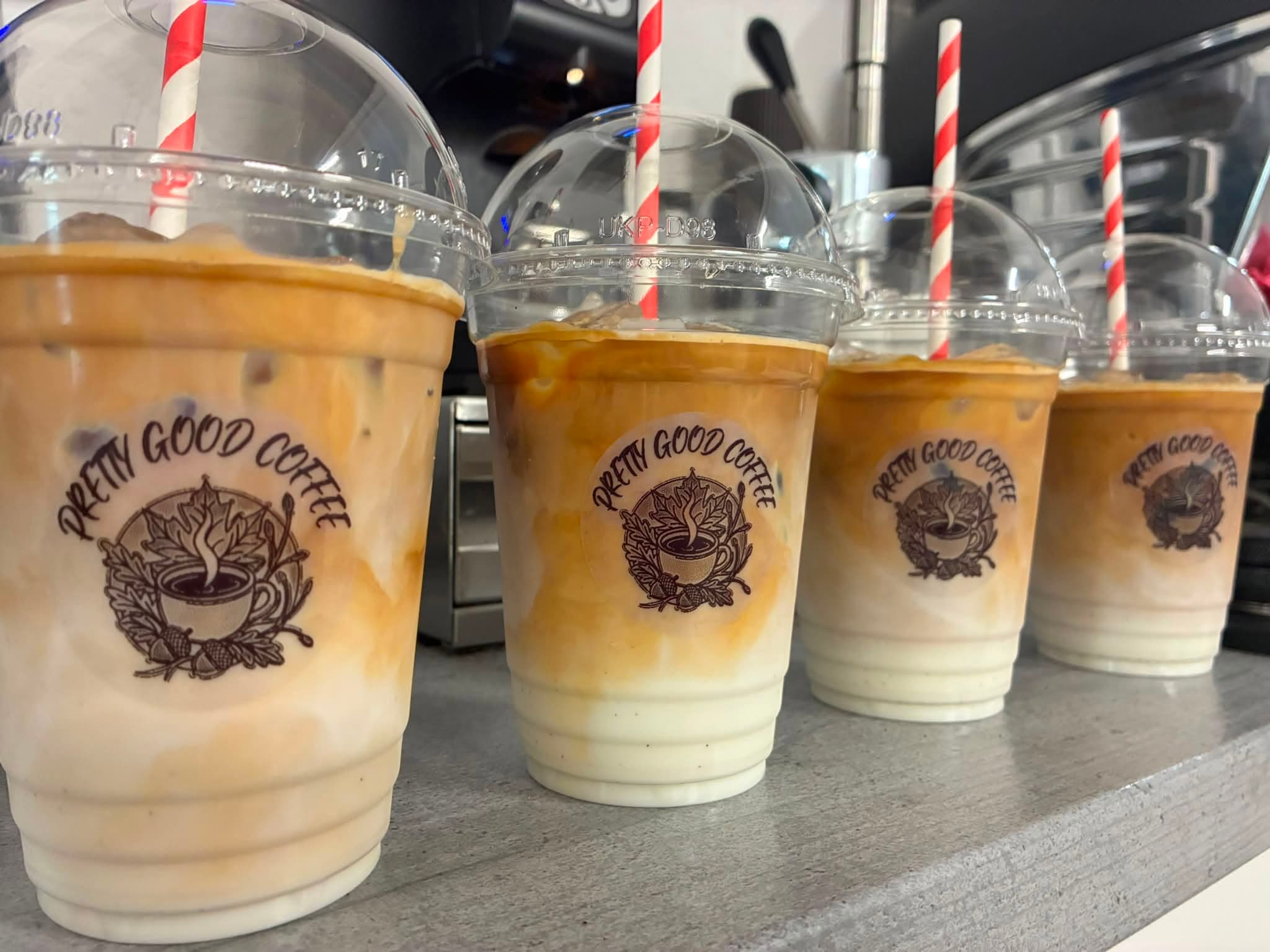 Four iced lattes in Pretty Good Coffee branded cups, lined up on the counter